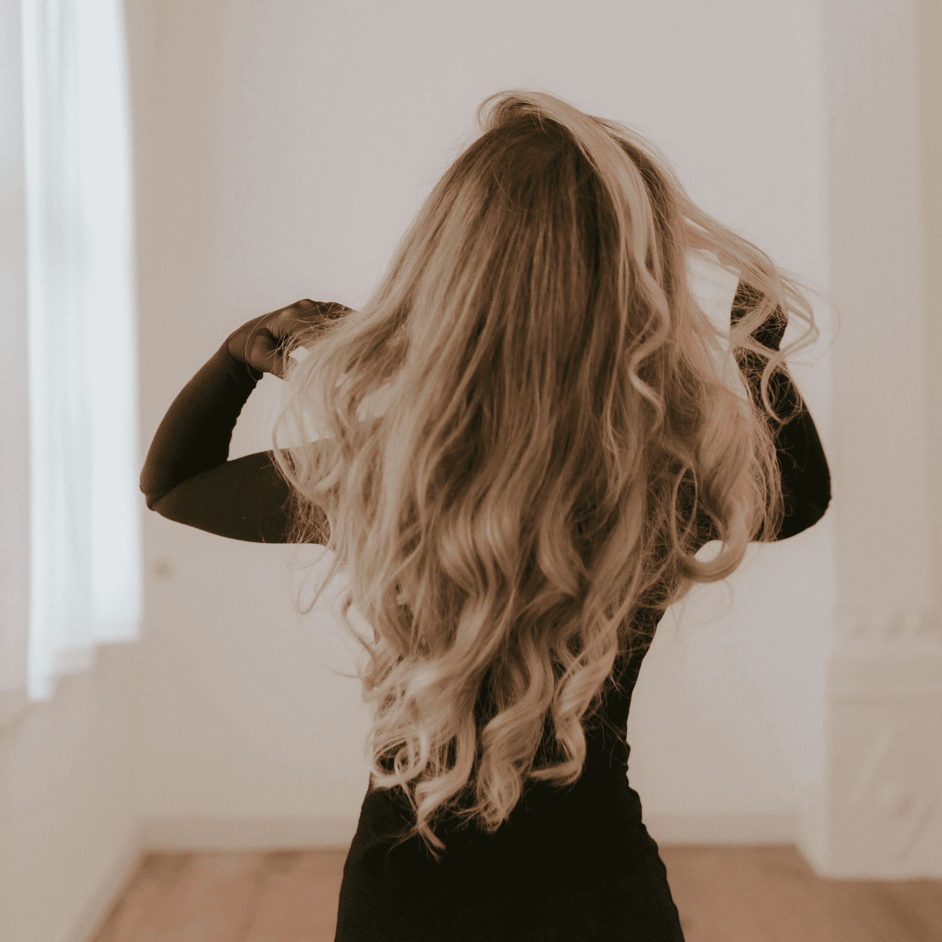 Blonde woman with long, wavy hair facing away in a bright, minimalist room.