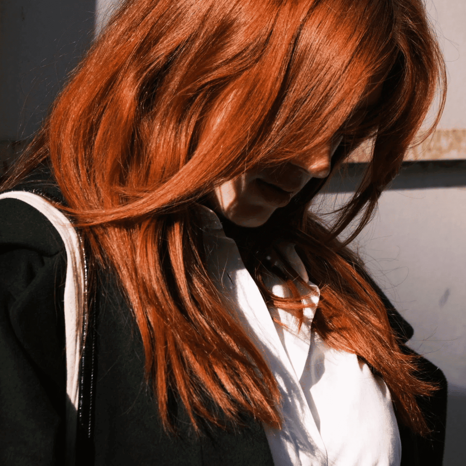 Red-haired woman in a black coat, standing in partial shadow against a textured wall.