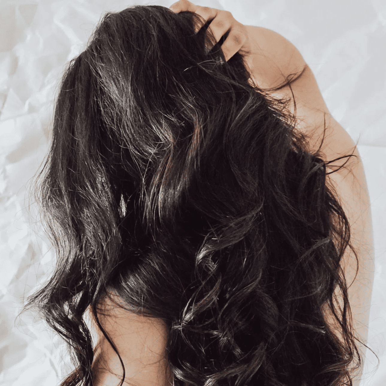 Long, wavy dark hair cascading over a person's back, against a crumpled white background.